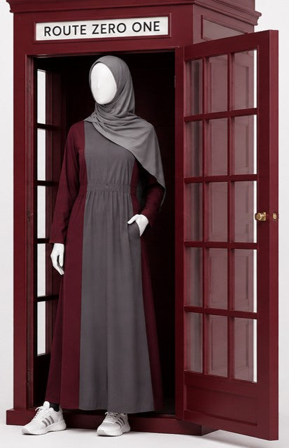 A grey and burgundy modest dress with a grey hijab in a red telephone booth